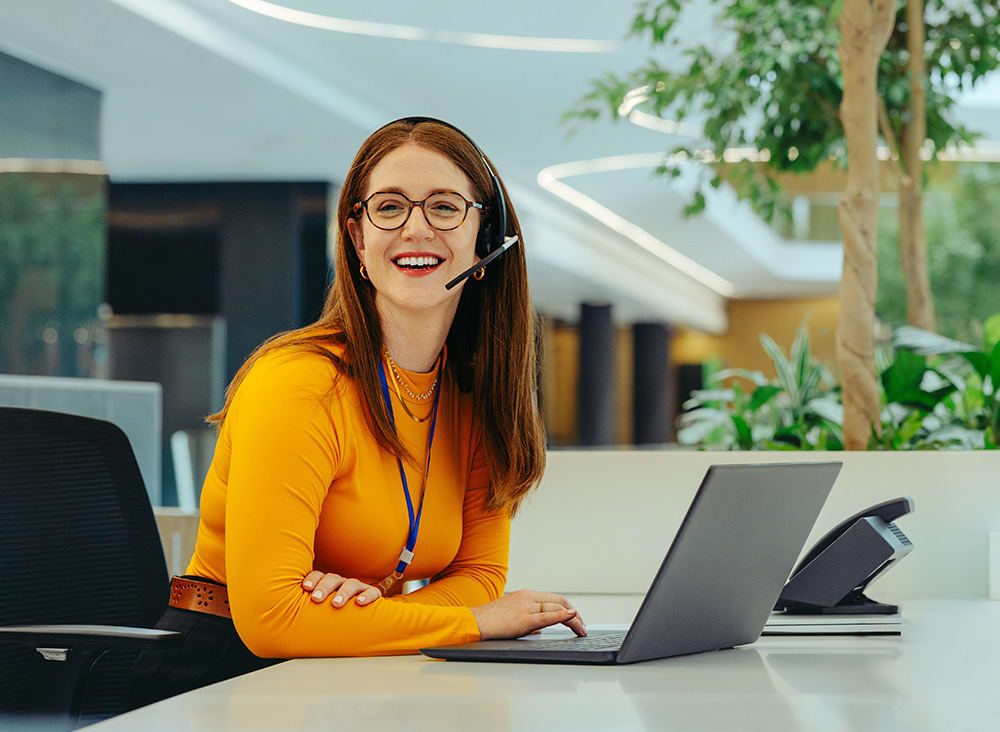 Person sitting at a desk in an office, wearing a yellow top and headset, looking at a laptop. An office phone is on the desk, with plants in the background.