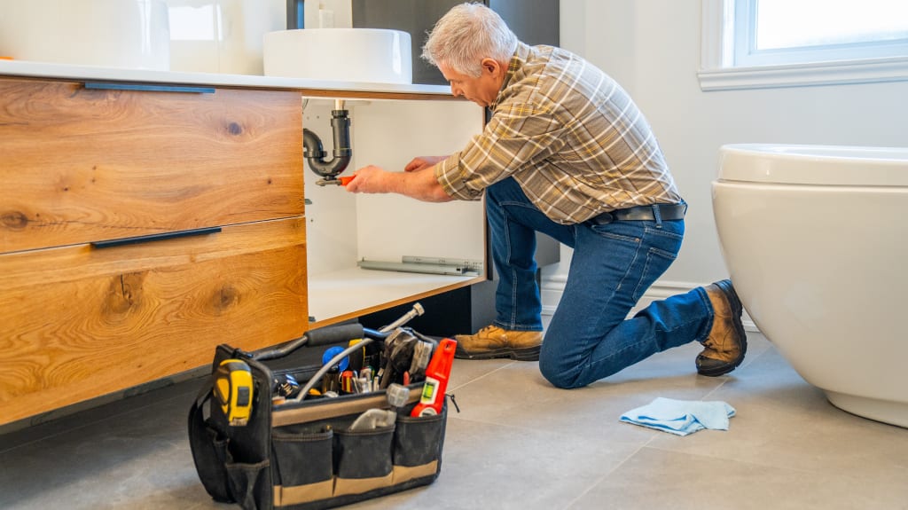 Un entrepreneur spécialisé en plomberie procède à des travaux sur la tuyauterie du lavabo d’une salle de bain, dans le cadre du service TravauxExpress.