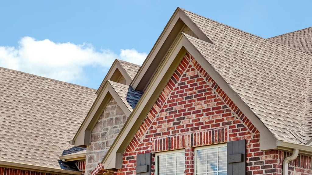 Toits en pente recouverts de bardeaux, avec des murs en briques rouges et pierres décoratives, fenêtres à volets noirs, sous un ciel bleu clair avec quelques nuages.