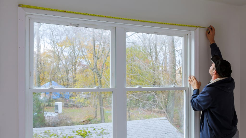 A person measures a large window opening with a tape measure before installation or replacement.