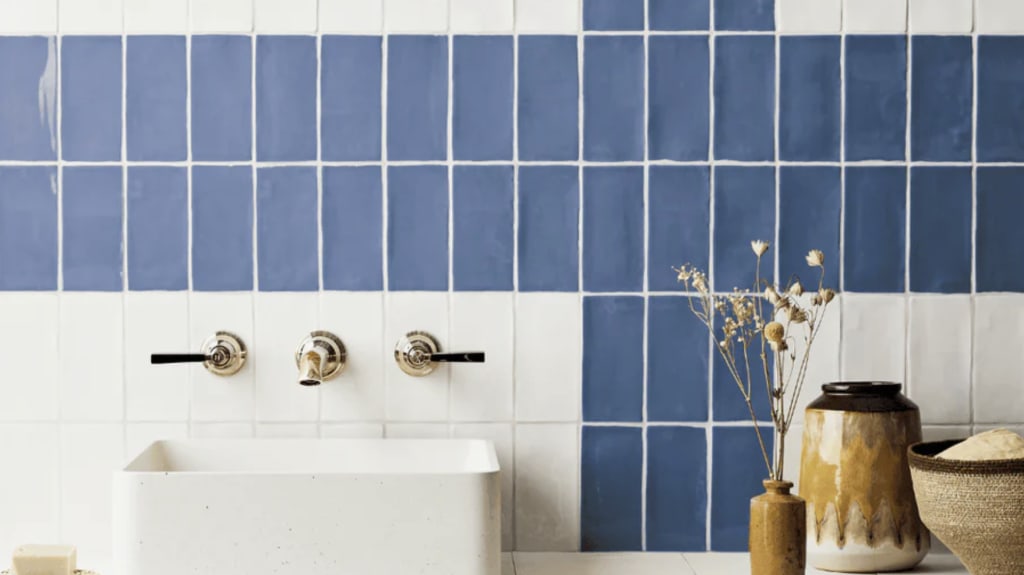 Dans une salle de bain, vasque blanche posée sur un meuble en bois brut recouvert de tuiles blanches, mur garni de tuiles rectangulaires bleu denim et blanc alignées à la verticale, pots en céramique à glaçure