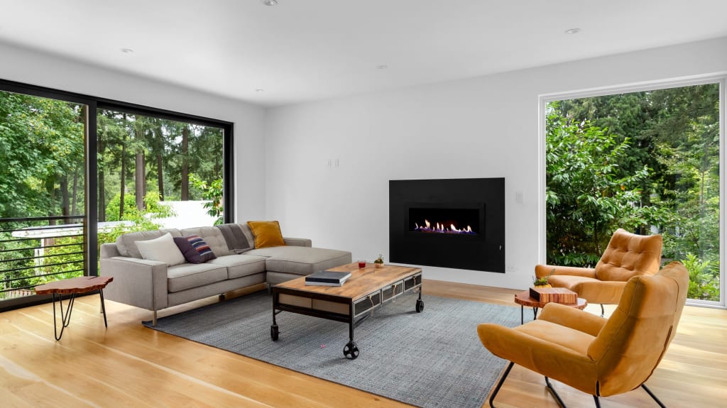 Living room decorated with a grey sofa and two ocre brown leather armchairs, built-in fireplace, and large bay windows overlooking a lush forest