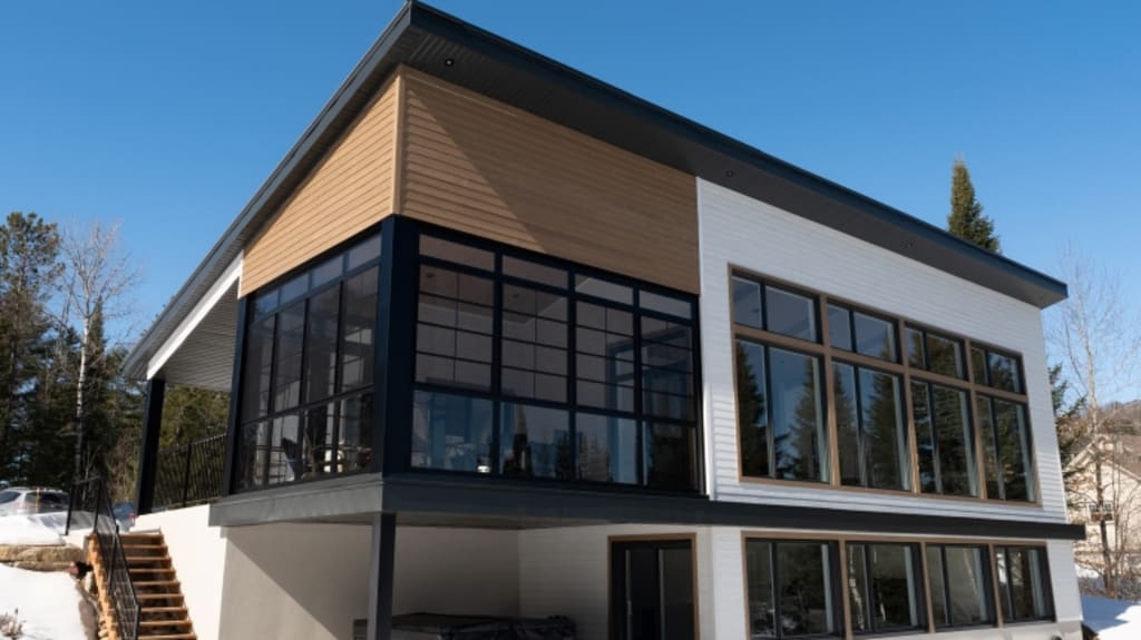 Modern two-storey house on sloped lot with black glass porch, brown and white engineered wood exterior cladding, lakefront and black pitched metal roof.