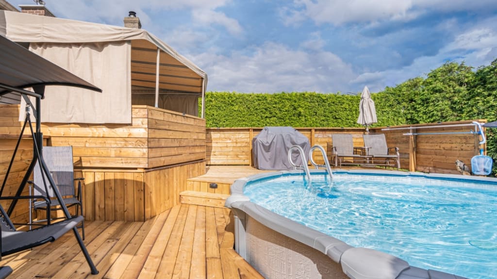 Three-level terrace made with treated wood decorated with a beige awning, chairs, a swing, and a BBQ, surrounding an above-ground pool.