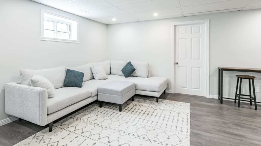 Bright basement with large white sectional, rug with a geometric pattern, and wooden bench