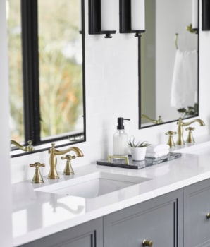 Miroirs aux contours noirs et robinetterie dorée dans une salle de bain avec tiroirs gris