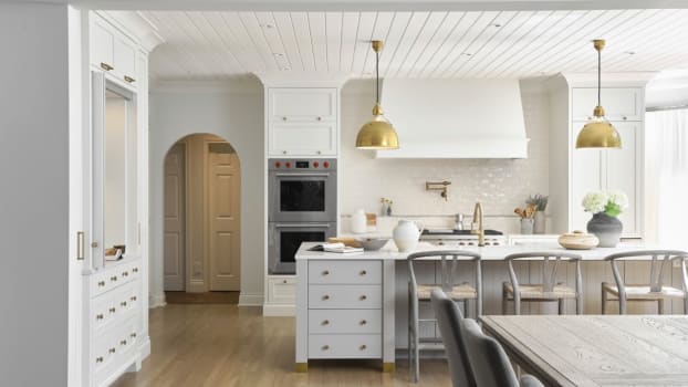 Wooden ceiling laths, gilded metal fixtures, plaster hood, arched entrance and white furniture in a seaside kitchen