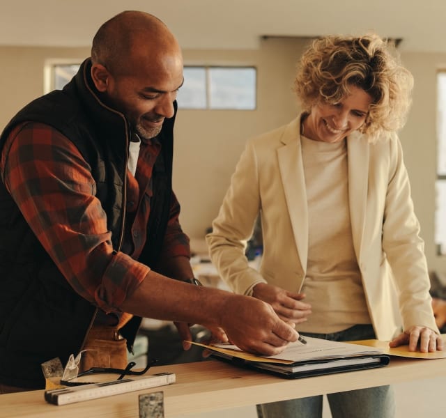 Deux personnes discutent autour d’une table en bois dans une pièce en rénovation, avec des outils et un carnet ouvert. Elles semblent examiner des plans ou des documents liés au projet.