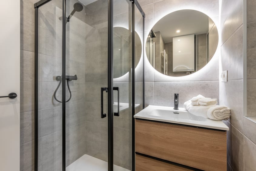 Douche préfabriquée dans le coin d’une petite salle de bain avec meuble vanité et miroir rond éclairé