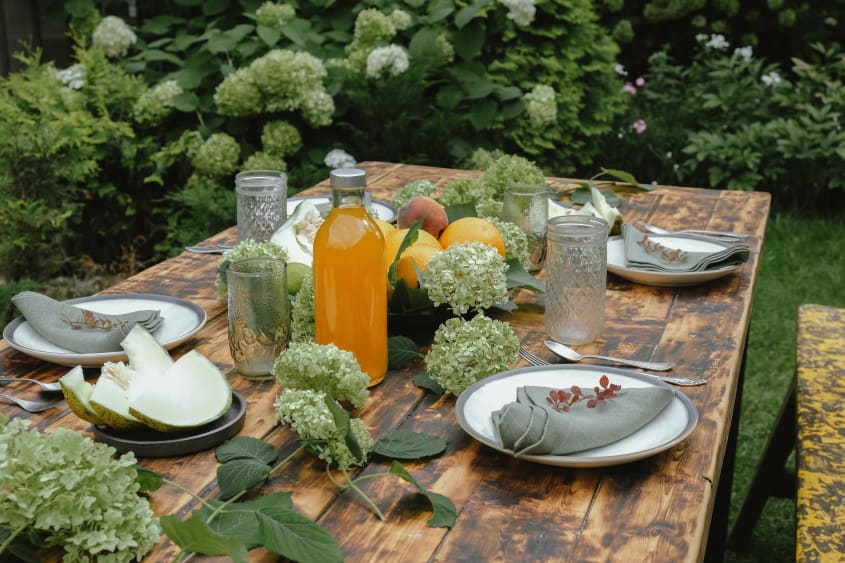 Close-up view of rustic wooden table with dinner place settings, fruit, orange juice bottle and pale green hortensia