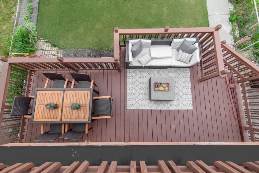 Top-down view of a small wooden brown L-shaped deck, dining set and couch covered with gray and white cushions