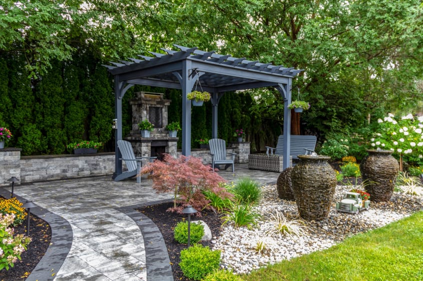 Surrounded by nature, a dark gray pergola overtops a stone patio, fireplace and wood furniture