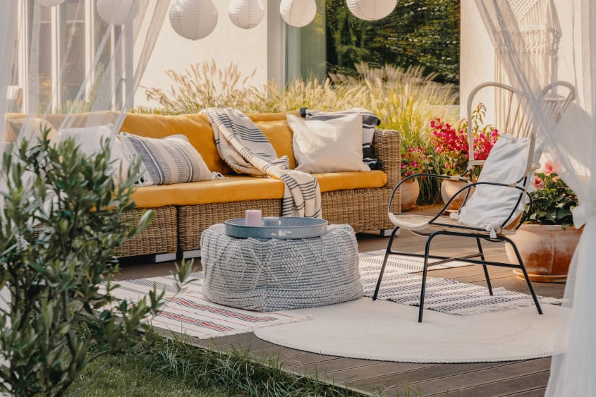 Boho style deck with wicker couch coverd with yellow, white and navy cushions, armchair, pouf used as a table and sheer curtains