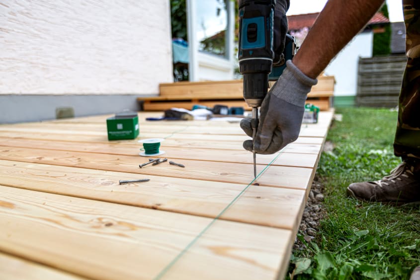 Un entrepreneur en construction visse une planche de mélèze pour l’installation d’une terrasse extérieure.