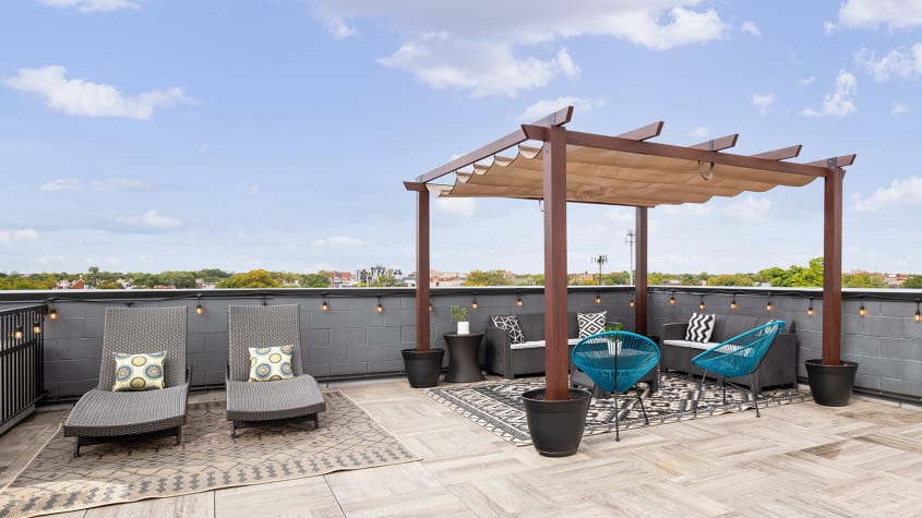 Terrasse moderne avec pergola en bois, chaises longues grises et fauteuils bleus, décorée de coussins et pots noirs, sous un ciel bleu avec quelques nuages, vue sur des arbres en arrière-plan.