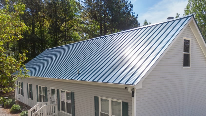 Maison blanche avec toit en métal bleu à pans inclinés, entourée d’arbres verts sous un ciel clair, avec plusieurs fenêtres et une petite entrée à l’avant.