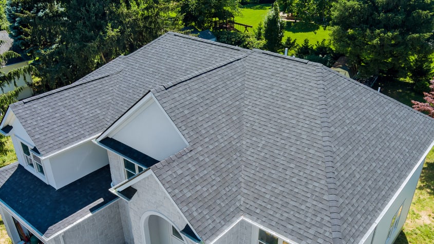 Vue aérienne d’un toit en bardeaux gris foncé avec plusieurs pentes, sur une maison aux murs clairs et détails en pierre, entourée d’arbres verts et d’un jardin en arrière-plan.