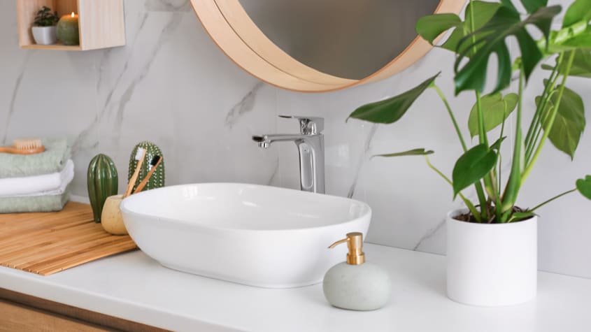 Lavabo blanc moderne avec robinet chromé, miroir rond en bois, plante verte et accessoires déco sur comptoir blanc