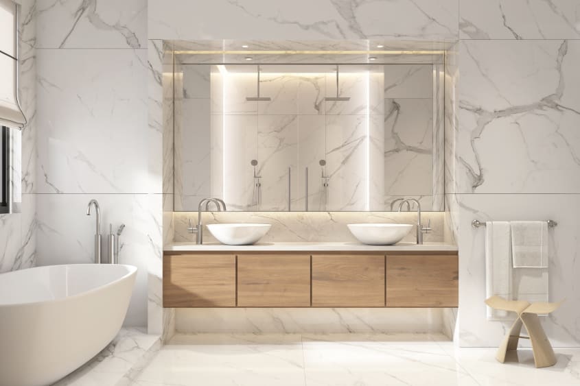 Vaste salle de bain aux murs et plancher recouverts de dalles grand format de marbre blanc veiné de gris, baignoire autoportante, grand miroir mural installé dans une alcôve, meuble de rangement flottant à quatre portes en bois, avec deux lavabos vasques 