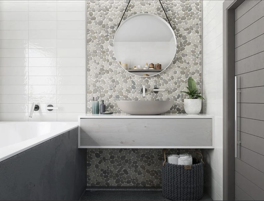 Salle de bain avec mur recouvert de tuiles céramiques blanches et de galets plats de plusieurs teintes de gris. Baignoire encastrée, comptoir flottant sur lequel reposent une vasque en porcelaine, des accessoires beauté et une plante
