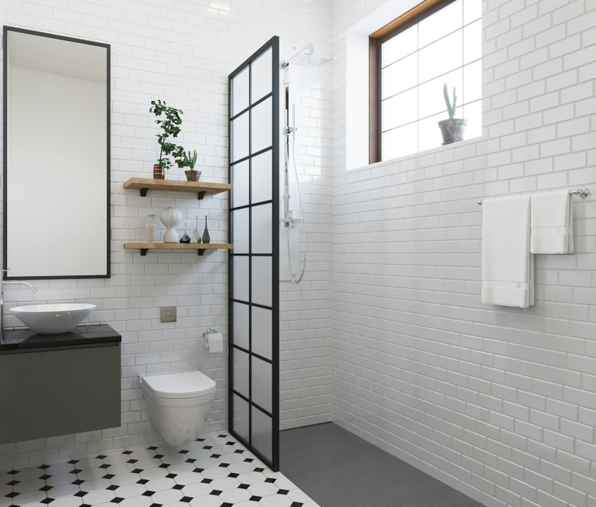Dans une salle de bain blanche lumineuse, une douche à l’italienne en coin, séparée par une cloison verrière, murs couverts de tuiles céramiques façon métro, plancher à carrelage cabochon, long miroir rectangulaire, étagères en bois avec flacons et plante