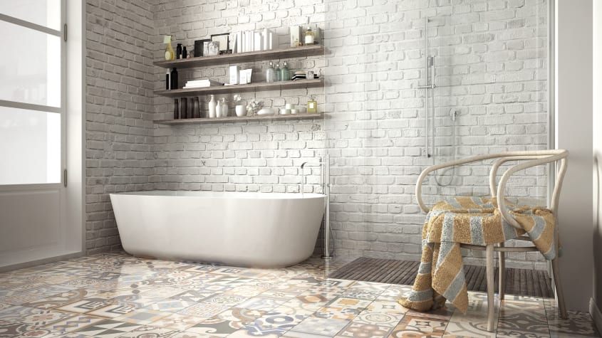 Salle de bain avec mur en briques peintes en gris pâle, trois étagères en bois avec produits de beauté et livres, baignoire autoportante, douche en coin, plancher couvert d’un carrelage en ciment patchwork, chaise Thonet en bois courbé, serviette éponge r
