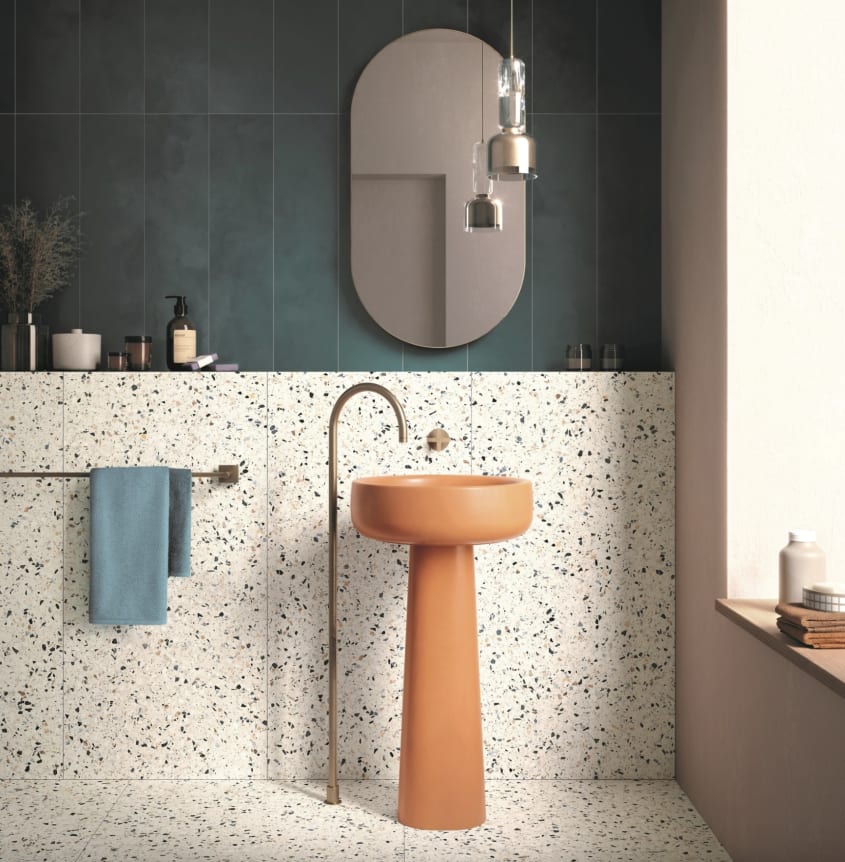 Dans une salle de bain de style Memphis, un lavabo sur colonne orange cantaloup avec robinet en laiton fixé au sol, demi-mur plein et plancher décorés de carrelage en terrazzo, partie supérieure du mur recouverte de longs zelliges vert sapin, miroir ovale