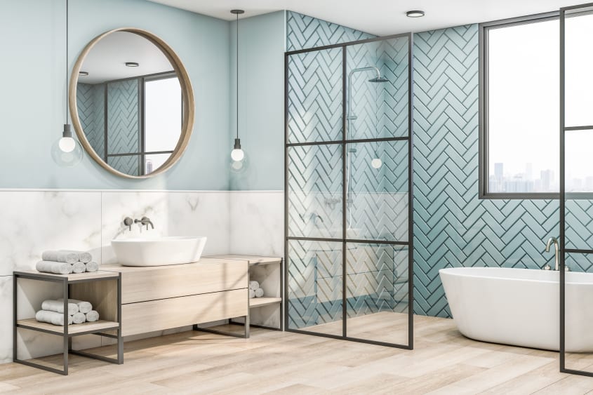 Vaste salle de bain avec espace de la douche à l’italienne et de baignoire ovale délimité par une cloison verrière, mur couvert de zelliges bleu mentholé, grand miroir rond encadré de bois, suspensions avec ampoules globes, meuble de rangement en bois cla