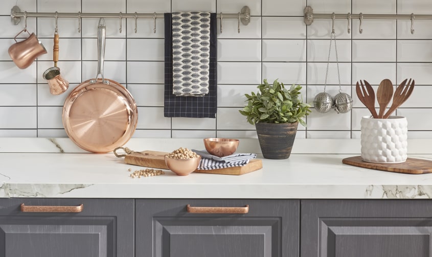 Dans une cuisine farmhouse, un dosseret en carrelage métro blanc, un comptoir marbré sur des armoires Shaker grises et plusieurs accessoires pour cuisiner comme un poêlon, des saucières et des bols en cuivre, des ustensiles en bois dans un pot et un bouqu