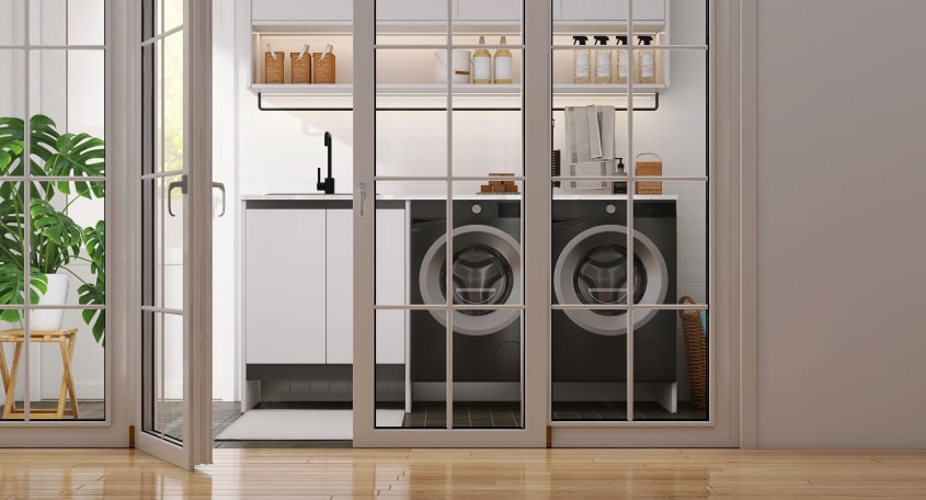 Portes françaises coulissantes à cadre blanc, ouvertes sur une salle de lavage avec deux laveuses noires, lavabo, étagères avec produits ménagers et plante monstera