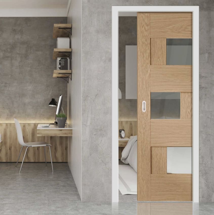 Porte coulissante escamotable ornée de trois fenêtres, donnant accès à une chambre à coucher. De l’autre côté du mur séparateur, un espace de travail avec table flottante et chaise en plastique moulé
