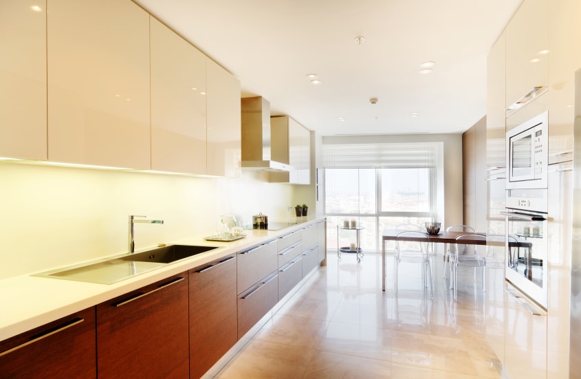 Cuisine moderne aménagée en longueur avec armoires minimalistes de couleur blanc crème et bois, plancher en marbre, table avec quatre chaises design en plastique transparent