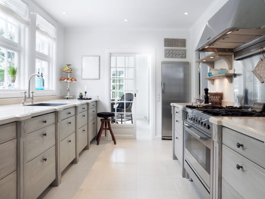 Grande cuisine de style campagnard aménagée en longueur avec armoires grises, réfrigérateur, cuisinière et hotte en acier inoxydable, et des portes françaises