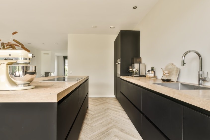 Cuisine aménagée le long d’un mur, armoires inférieures noires avec comptoirs en travertin, plancher en bois, îlot avec un batteur sur socle
