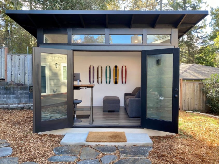 Bureau maison installé dans un studio de jardin à porte double vitrée, table de style établi, canapé et ottoman, planches à roulettes accrochées sur le mur