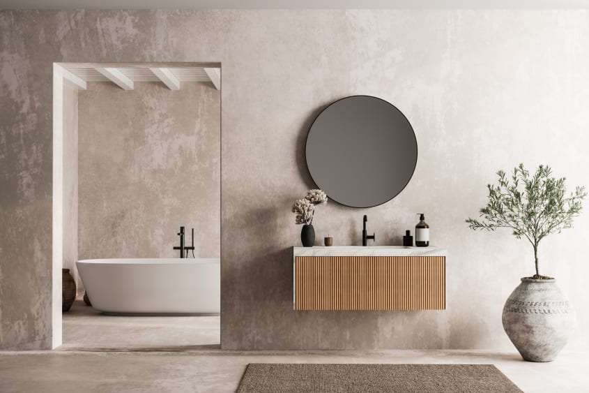 Salle de bain spacieuse et luxueuse à murs et plancher en ciment brut, plafond blanc en lambris et poutres. Dans une pièce, meuble-lavabo suspendu à panneau cannelé et comptoir en marbre sur lequel sont déposés un vase à fleurs, une bougie, un flacon-pomp