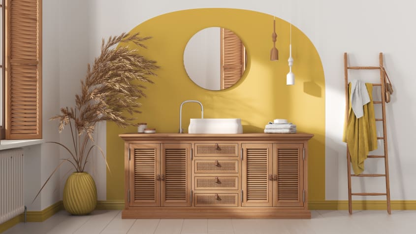 Dans une salle de bain de luxe, un meuble de rangement en bois avec portes en louvres et tiroirs en cannage, vasque blanche, miroir rond sur un mur d’accent jaune et échelle servant de porte-serviettes