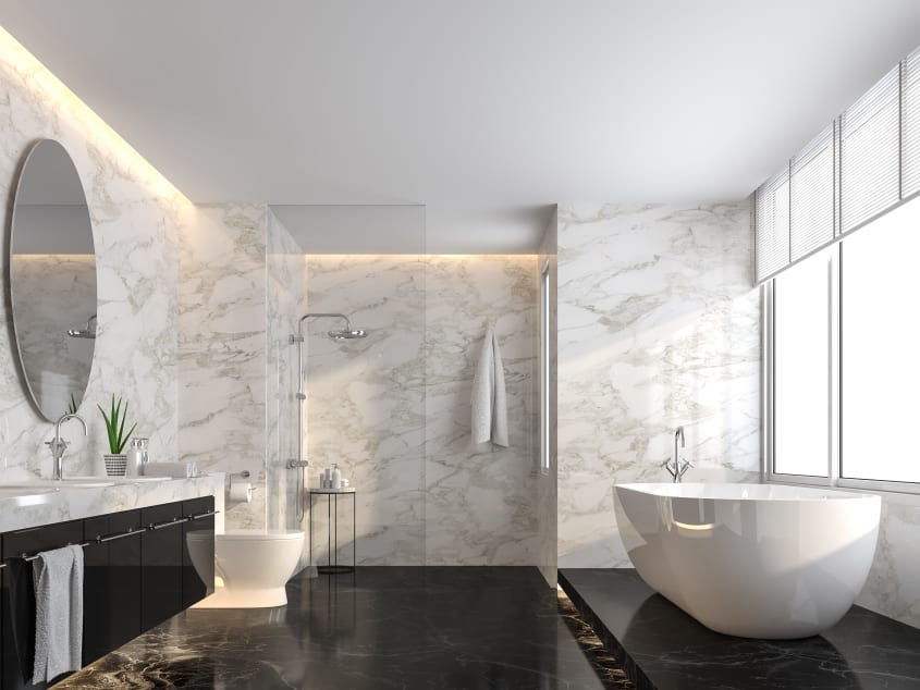 Salle de bain luxueuse très lumineuse, avec murs couverts de marbre blanc à veinures grises. La baignoire et la toilette en porcelaine blanche contrastent avec le plancher et le meuble-lavabo noirs. Une paroi de verre délimite la douche à l’italienne au f