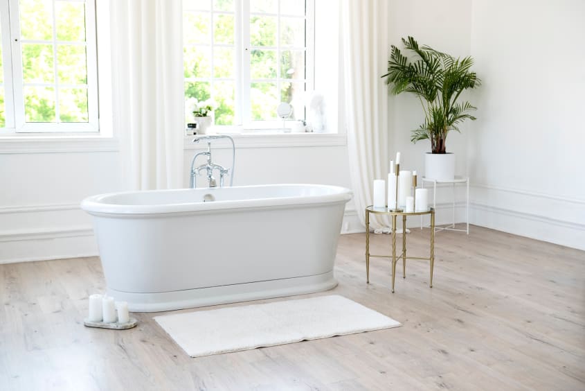 In an all-white bathroom with windows overlooking a green landscape, a freestanding oval bathtub with silver hardware, candles placed on the light wood floor and on a side shelf