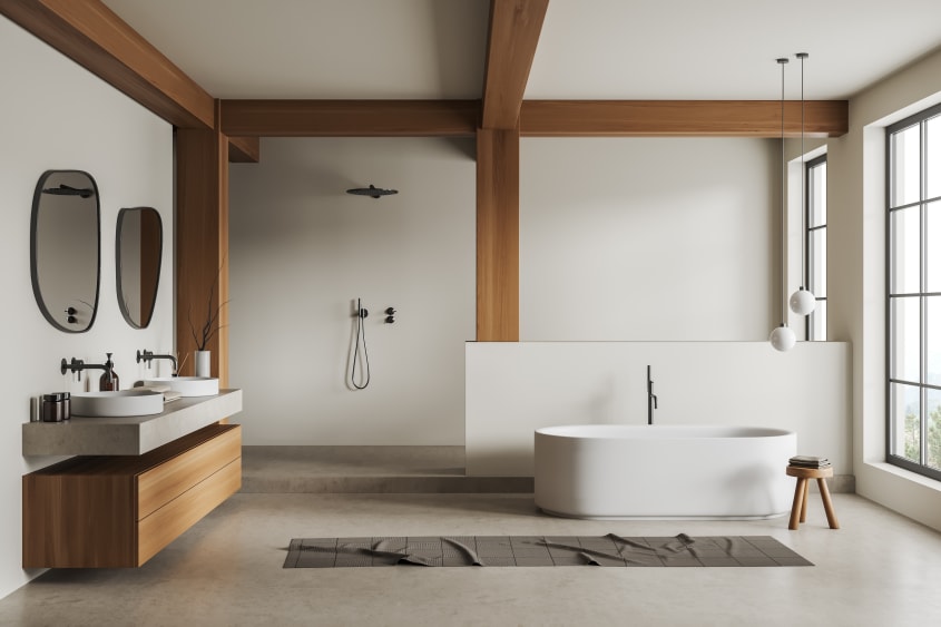 Salle de bain blanche de grande dimension, décorée de poutres et d’un meuble de rangement suspendu en bois, deux vasques en porcelaine, deux miroirs aux formes organiques, baignoire ovale autoportante, tabouret de bois, tapis de bain et fenêtres à carreau