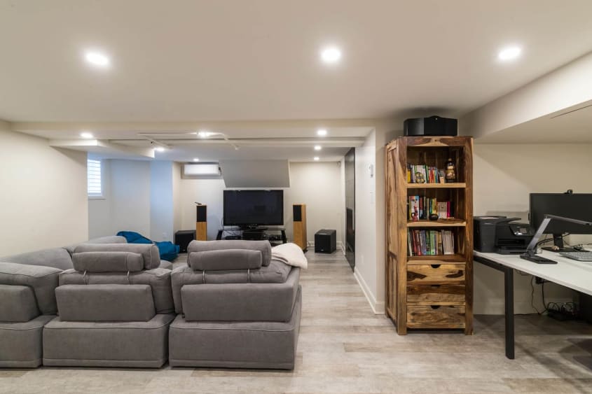Salle familiale d'un sous-sol rénovée avec divan gris, télé, coin bureau, murs beiges et étagère en bois