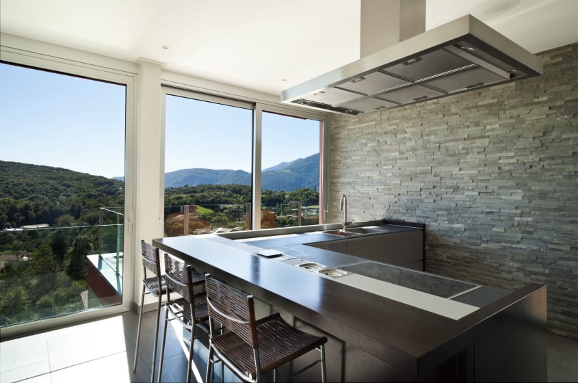 Picture windows with mountain views in a modern kitchen with gray brick wall and L-shaped island