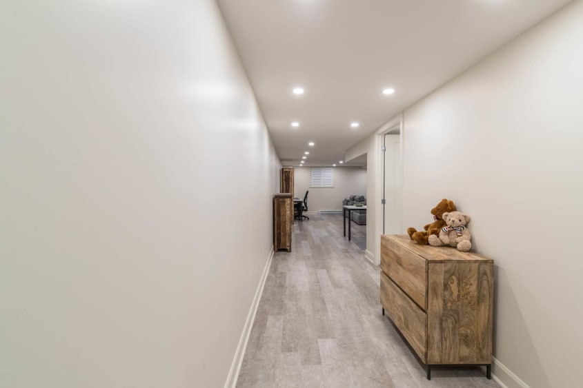 Corridor d'un sous-sol moderne rénovée avec murs sable, meuble en bois, plancher flottant gris pâle et éclairage encastré