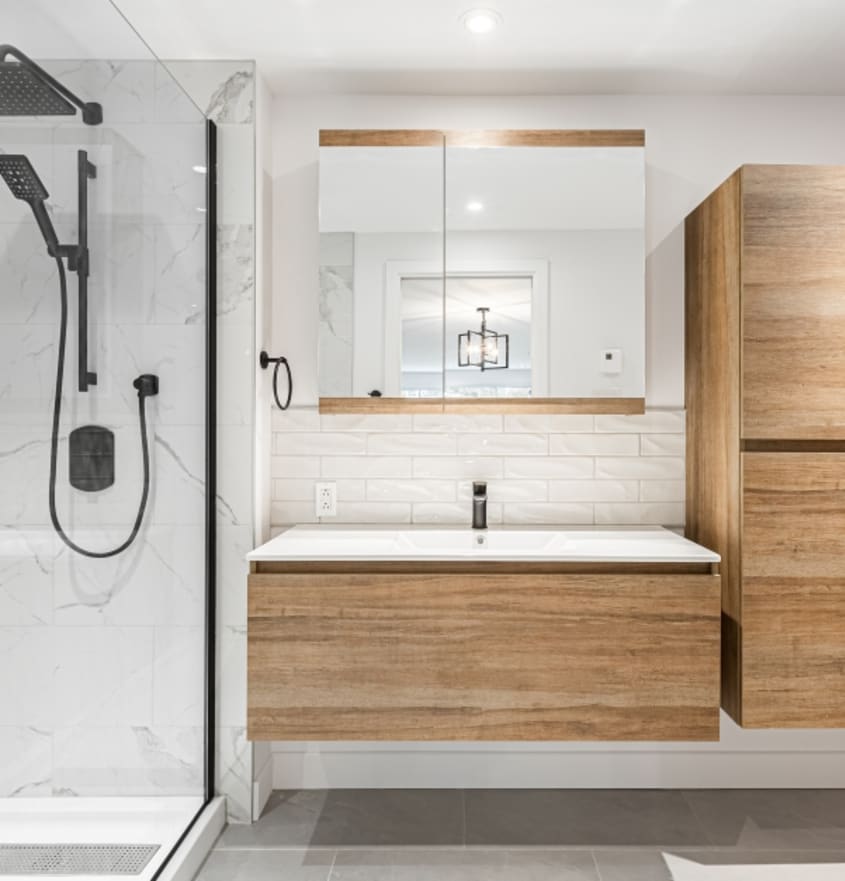 Modern white bathroom with floating vanity and brown cabinets, white sink, white “metro” style ceramic backsplash with corner glass shower, black faucets, and grey ceramic floor.