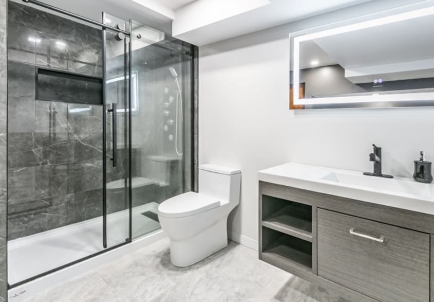 Salle de bain moderne dans les teintes de gris et blanc avec grande douche comprenant porte vitrée et mur de céramique grise, miroir intelligent sur mur blanc, meuble-lavabo gris de forme rectangulaire avec rangements et robinetterie foncée ainsi plancher