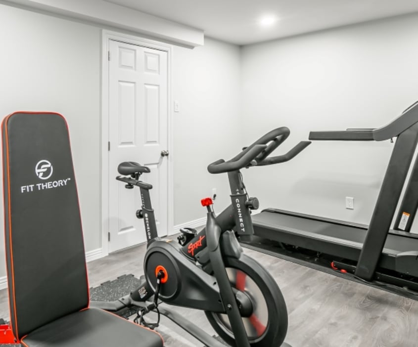 Bel aménagement d’une salle de gym sobre et lumineuse avec mur et plafond blanc, plancher flottant gris comprenant vélo stationnaire, banc pour musculation ainsi que tapid roulant.