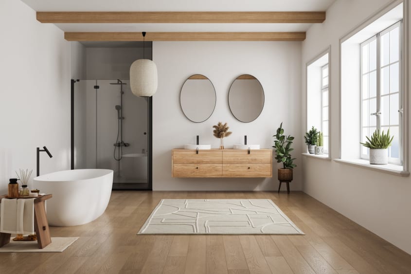 Salle de bain lumineuse de style scandinave, à plancher, meuble-lavabo et poutres en bois pâle, fenêtres à carreaux, deux miroirs ovales, baignoire autoportante nlanche, tapis de bain nervuré, tabouret bas en bois avec produits de soins, lampe lanterne en